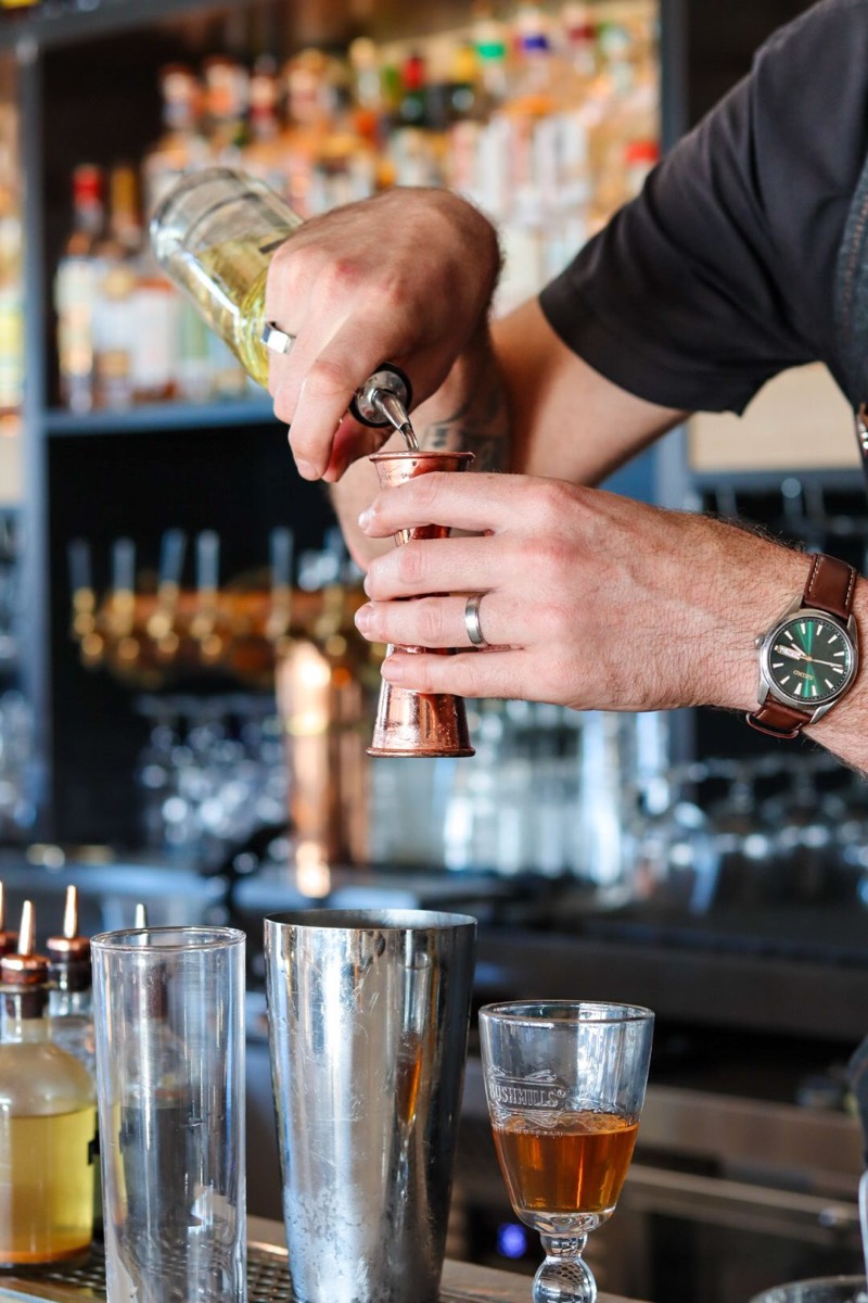 A bartender pouring spirits into a copper jigger over a lineup of shakers and glasses at the back bar