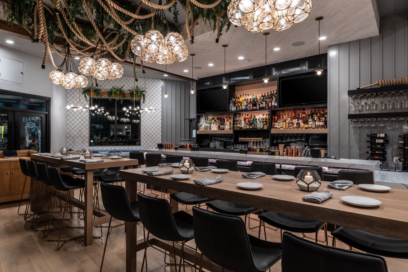 Warmly lit dining tables set with plates beneath clusters of globe chandeliers wrapped in rope and greenery